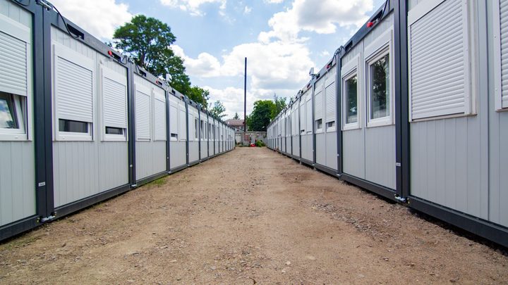 Duas fileiras longas de módulos em container cinza, com janelas de persiana branca fechada, assentados sobre pés metálicos dos dois lados de um caminho de terra batida.