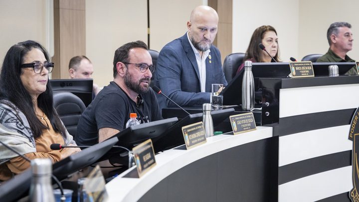 Vereadores de Itapema sentados na bancada curva do plenário durante uma sessão, com microfones de haste, monitores e placas de identificação douradas à frente de cada lugar.