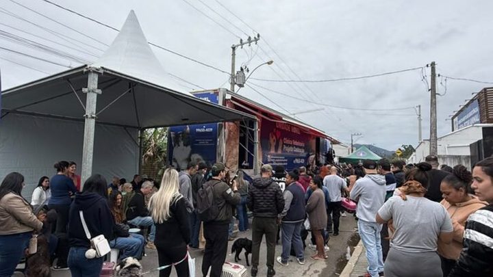 Fila de tutores com c&atilde;es na guia e caixas de transporte numa rua de Itapema, ao lado de uma tenda branca e do caminh&atilde;o da unidade m&oacute;vel de castra&ccedil;&atilde;o estacionado junto ao meio-fio.