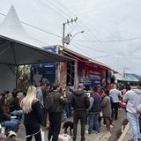 Fila de tutores com cães na guia e caixas de transporte numa rua de Itapema, ao lado de uma tenda branca e do caminhão da unidade móvel de castração estacionado junto ao meio-fio.