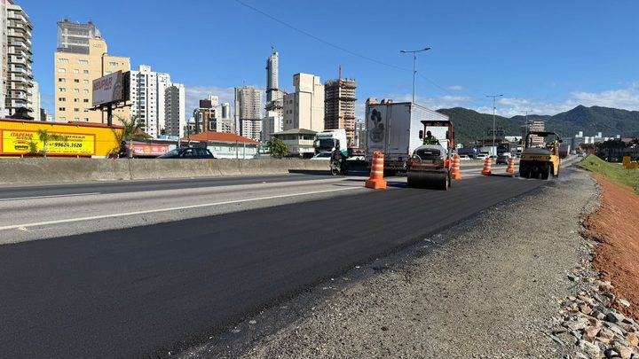 Pista da marginal da BR-101 em Itapema recém-asfaltada, com rolo compactador laranja e caminhão trabalhando ao fundo, cones de sinalização na faixa ao lado, prédios da cidade no horizonte e brita solta na borda direita da pista.