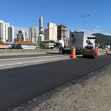Pista da marginal da BR-101 em Itapema recém-asfaltada, com rolo compactador laranja e caminhão trabalhando ao fundo, cones de sinalização na faixa ao lado, prédios da cidade no horizonte e brita solta na borda direita da pista.
