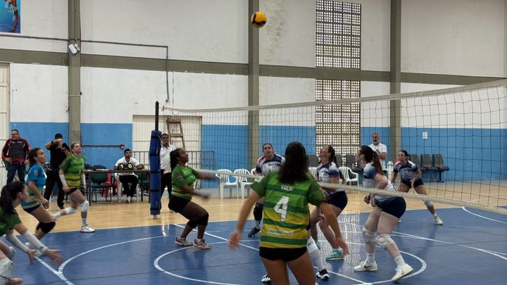 Partida de voleibol feminino em ginásio, com a bola no alto sobre a rede e jogadoras de duas equipes posicionadas em quadra azul.