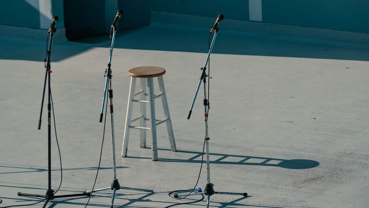 Palco vazio ao ar livre, com três pedestais de microfone montados sobre piso de concreto claro, um banquinho alto de madeira no meio e cabos soltos no chão, antes de a apresentação começar.