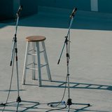 Palco vazio ao ar livre, com três pedestais de microfone montados sobre piso de concreto claro, um banquinho alto de madeira no meio e cabos soltos no chão, antes de a apresentação começar.