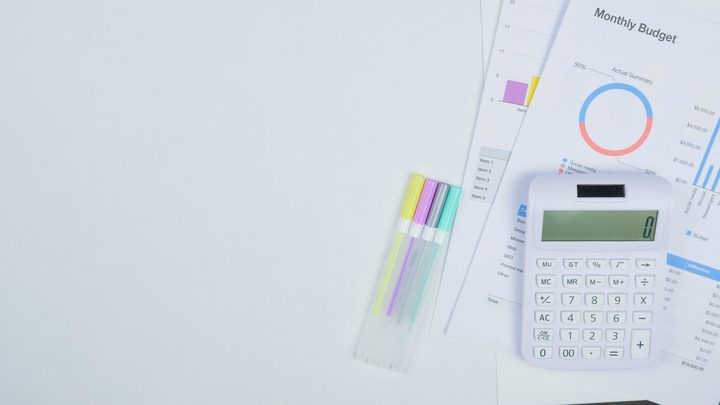 Mesa branca vista de cima com uma calculadora de teclas claras apoiada sobre folhas impressas de planilha de orçamento, com gráfico de rosca colorido e colunas de valores, e quatro canetas marca-texto alinhadas ao lado.