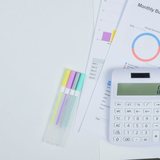 Mesa branca vista de cima com uma calculadora de teclas claras apoiada sobre folhas impressas de planilha de orçamento, com gráfico de rosca colorido e colunas de valores, e quatro canetas marca-texto alinhadas ao lado.