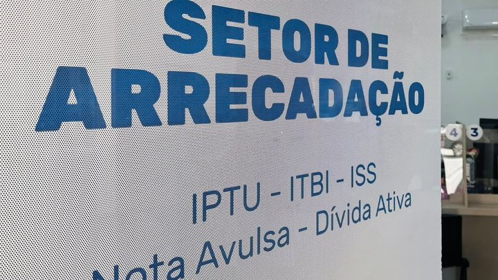 Placa adesivada no vidro do Setor de Arrecadação da Prefeitura de Itapema, com as palavras SETOR DE ARRECADAÇÃO em letras azuis grandes e, abaixo, a linha IPTU, ITBI, ISS, Nota Avulsa e Dívida Ativa.