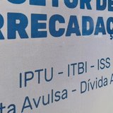 Placa adesivada no vidro do Setor de Arrecadação da Prefeitura de Itapema, com as palavras SETOR DE ARRECADAÇÃO em letras azuis grandes e, abaixo, a linha IPTU, ITBI, ISS, Nota Avulsa e Dívida Ativa.