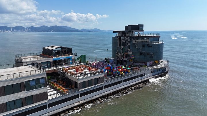 Vista aérea do píer da Foz do Rio Perequê, em Itapema, com prédio circular de vidro escuro na ponta, a guitarra do Hard Rock Cafe na fachada, terraços com mesas e o mar em volta.
