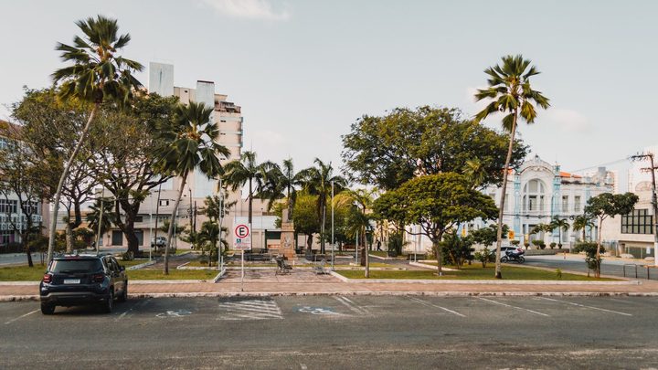 Rua larga de cidade litorânea com vagas de estacionamento demarcadas e vazias no primeiro plano, um carro estacionado à esquerda, placa de estacionamento regulamentado no canteiro e coqueiros diante de prédios ao fundo.