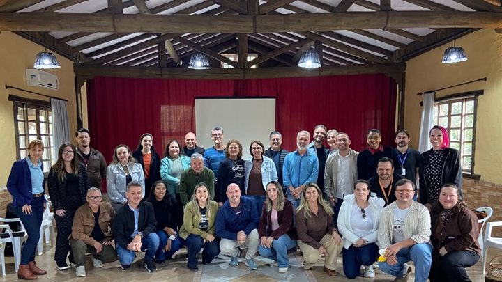 Cerca de trinta pessoas adultas posam em pé e agachadas para foto de grupo dentro de um salão de telhado aparente de madeira, com cortina vermelha e tela de projeção ao fundo.