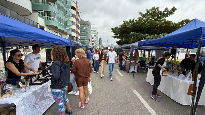 Tendas azuis montadas nos dois lados da avenida da orla de Itapema, com bancas de artesanato, visitantes circulando pelo meio da via e a linha de prédios da praia ao fundo.