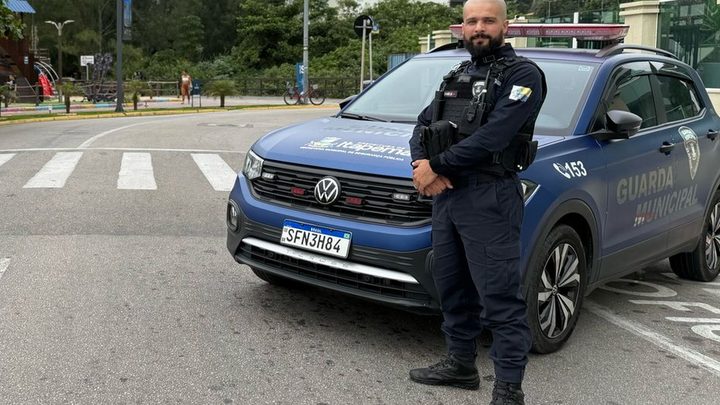 Agente da Guarda Municipal de Itapema, de uniforme escuro e colete, em pé ao lado de uma viatura azul atravessada na pista de uma avenida da orla, com faixa de pedestres à frente e a rua vazia.