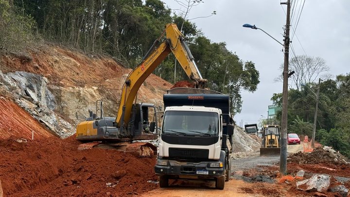 Escavadeira amarela com o braço estendido despejando terra vermelha na caçamba de um caminhão branco parado numa estrada de terra do Morro do Encano, com barranco recém-cortado e paredão de rocha exposta à esquerda e poste de energia à direita.