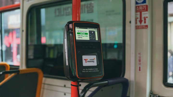 Validador de cartão preso a uma barra vermelha dentro de um ônibus urbano, com a tela acesa, a área de aproximação do cartão logo abaixo e as janelas do veículo desfocadas ao fundo.