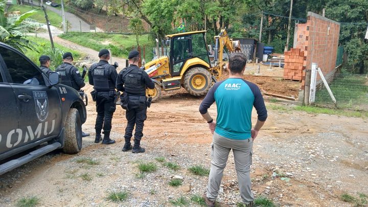 Retroescavadeira amarela parada em terreno de saibro ao lado de uma construção de tijolo sem reboco, com agentes da Guarda Municipal de costas junto a uma caminhonete e mata fechada ao fundo.