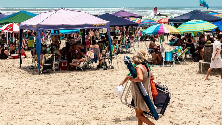 Praia cheia em dia de sol, com tendas e guarda-sóis coloridos ocupando a areia, cadeiras de praia espalhadas e o mar ao fundo.
