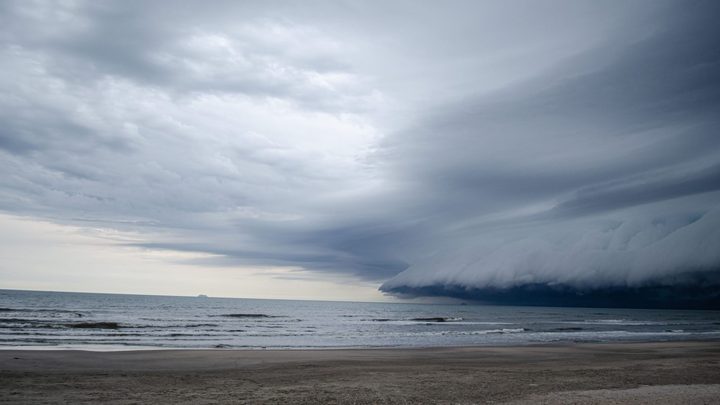 Nuvem escura e alongada de tempestade avançando sobre o mar em direção a uma faixa de areia vazia, com a água esverdeada e o céu claro ainda visível no lado esquerdo do horizonte.