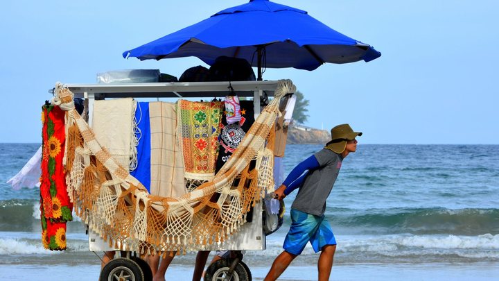 Ambulante de chapéu e camiseta cinza puxa pela areia um carrinho de duas rodas coberto por guarda-sol azul, carregado de redes de descanso, cangas e toalhas coloridas, com o mar quebrando ao fundo.