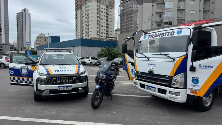 Caminhão plataforma branco com a palavra TRÂNSITO no para-brisa e faixas azuis e amarelas, ao lado de uma picape e de uma motocicleta com a mesma pintura, num pátio asfaltado com prédios altos de Itapema ao fundo.