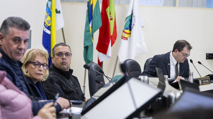 Vereadores de Itapema sentados lado a lado na bancada do plenário durante uma sessão, com microfones de haste e as bandeiras do Brasil e de Santa Catarina ao fundo.