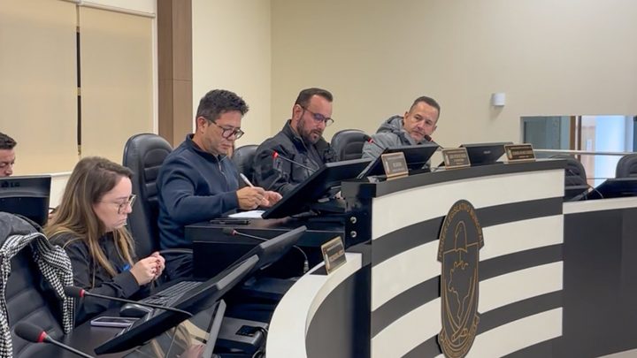 Quatro pessoas sentadas lado a lado na bancada preta e branca do plenário da Câmara de Itapema, com microfones de haste, monitores e placas de identificação douradas à frente, e o brasão do Poder Legislativo Municipal gravado na frente da bancada.