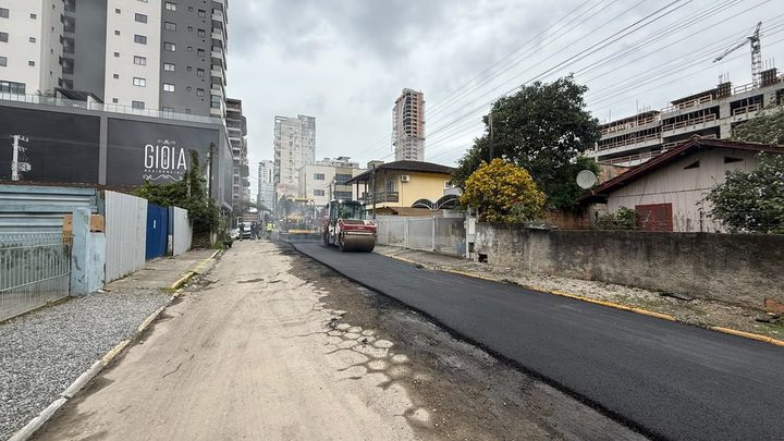 Rua 250 em obras na Meia Praia, em Itapema, com metade da pista já coberta por asfalto novo e escuro, a outra metade em terra batida com buracos, rolo compactador ao fundo e prédios altos dos dois lados.