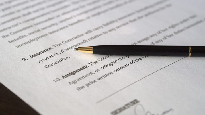 Close de um contrato impresso em papel branco, com as cláusulas numeradas em destaque e uma caneta esferográfica preta de detalhe dourado apoiada sobre o texto, ao lado da linha de assinatura.