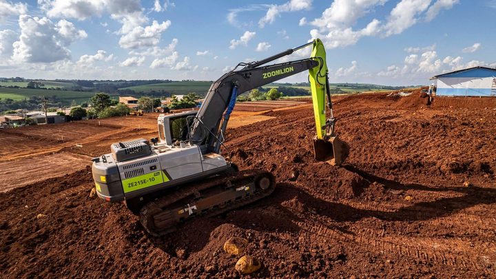 Escavadeira de esteira verde e cinza trabalhando sobre um terreno de terra vermelha revolvida, com a caçamba levantada, marcas de esteira no solo e área rural verde ao fundo sob céu azul.