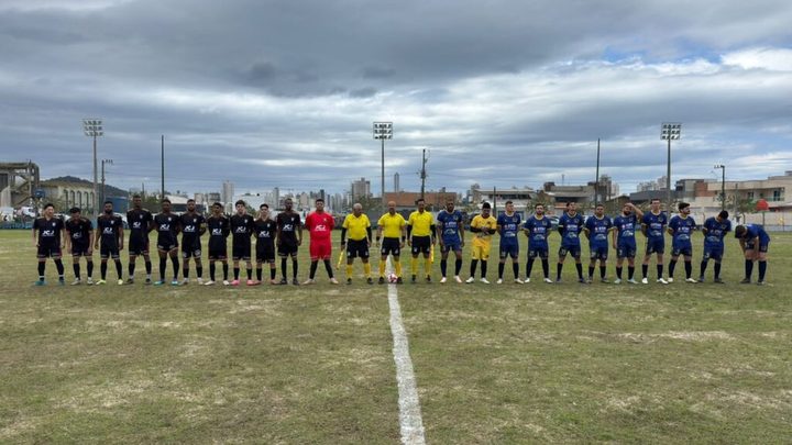 Duas equipes de futebol amador perfiladas lado a lado no meio do campo antes do apito inicial, uma de uniforme preto e outra de azul, com três árbitros de amarelo e a bola no círculo central, e prédios de Itapema ao fundo.