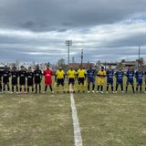 Duas equipes de futebol amador perfiladas lado a lado no meio do campo antes do apito inicial, com três árbitros de amarelo entre elas.