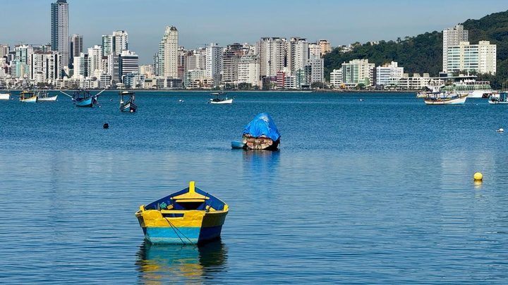 Barco de pesca pequeno, pintado de azul e amarelo, fundeado em água calma e esverdeada, com outros barcos ao fundo e a linha de prédios altos de Itapema atrás, sob céu azul.