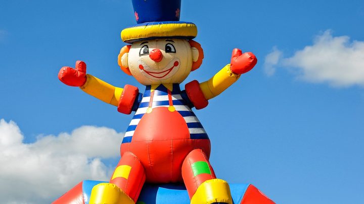 Brinquedo inflável gigante em forma de palhaço, com cartola azul, colete listrado e luvas vermelhas, inflado ao ar livre contra um céu azul com poucas nuvens.