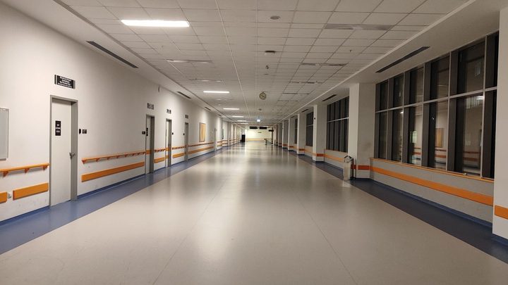 Corredor largo e vazio de hospital, com piso cinza brilhante, paredes brancas com corrimão laranja, portas fechadas à esquerda e janelas altas à direita.