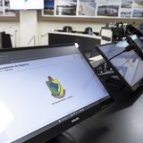 Monitor ligado na bancada do plenário da Câmara de Itapema mostrando a página do sistema da Casa, com um microfone de haste ao lado e cadeiras pretas vazias ao fundo.