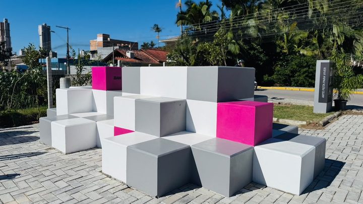 Banco público de cubos de concreto brancos, cinzas e rosa montado em degraus sobre calçada de paver clara, numa praça nova de Itapema, com coqueiros e casas ao fundo e um totem escuro à direita.