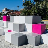 Banco público de cubos de concreto brancos, cinzas e rosa montado em degraus sobre calçada de paver clara, numa praça nova de Itapema, com coqueiros ao fundo.