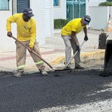 Dois trabalhadores de camiseta amarela espalhando massa asfáltica preta com rodos numa rua de Itapema, diante de um prédio branco, com a caçamba de uma pá-carregadeira despejando asfalto à direita.