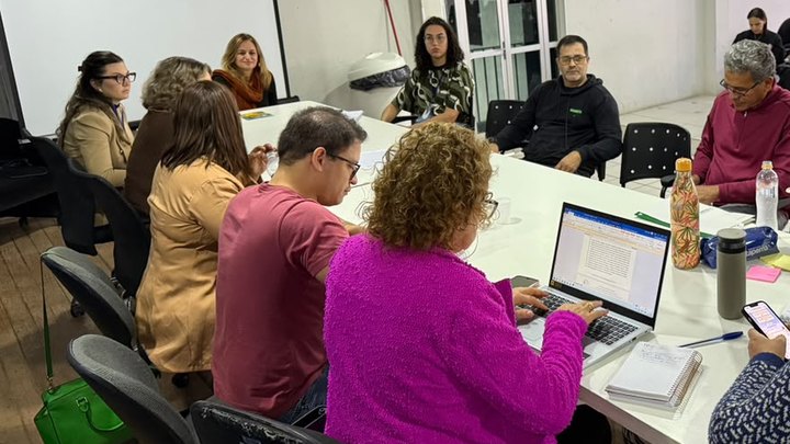 Reunião do conselho da criança e do adolescente de Itapema em volta de uma mesa branca comprida, com cerca de dez conselheiros sentados, um notebook aberto mostrando documento de texto, garrafas térmicas e caderno de anotações sobre a mesa.