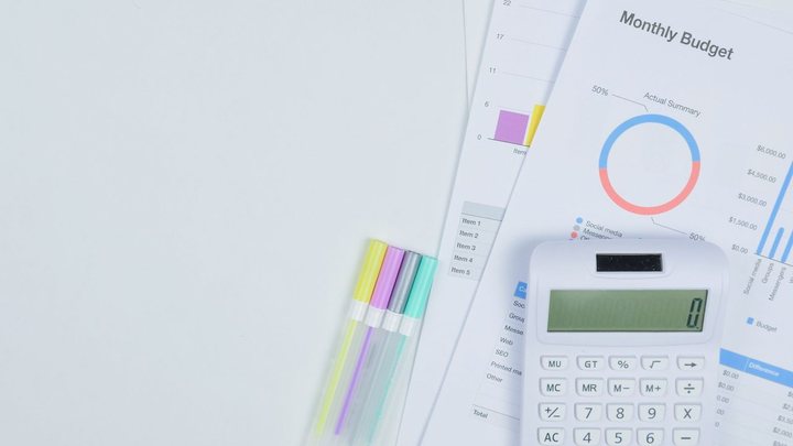 Mesa branca vista de cima com uma calculadora de bolso branca apoiada sobre folhas de planilha impressas, uma delas com o título Monthly Budget e um gráfico de rosca em azul e vermelho, ao lado de quatro canetas marca-texto coloridas.
