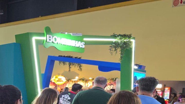 Pórtico verde de estande de feira de turismo, iluminado por fita de LED, com o nome Bombinhas em letras brancas sobre um recorte verde, trepadeira artificial pendurada na quina e um pórtico azul ao fundo.
