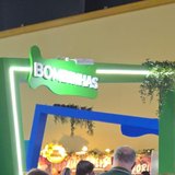 Pórtico verde de estande de feira de turismo, iluminado por fita de LED, com o nome Bombinhas em letras brancas sobre recorte verde e trepadeira artificial pendurada na quina.
