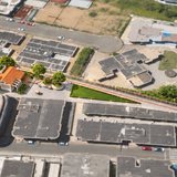 Vista aérea de um quarteirão do Centro de Itapema com o desenho do projeto da praça sobreposto à foto, mostrando um salão de telhado laranja, árvores novas e um passeio claro entre duas ruas.