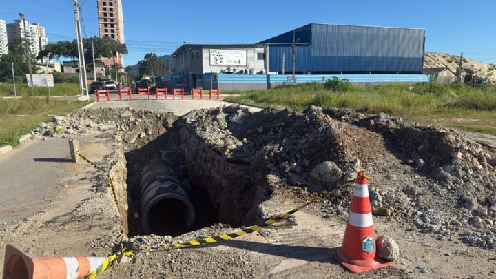 Vala aberta numa rua de terra de Itapema com um tubo grosso de concreto de drenagem assentado no fundo, montes de terra e pedra nas bordas, cones laranja e fita zebrada isolando o buraco, e um galpão azul ao fundo.