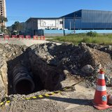 Vala aberta numa rua de terra de Itapema com tubo grosso de concreto de drenagem assentado no fundo, montes de terra nas bordas e cone laranja de sinalização ao lado.