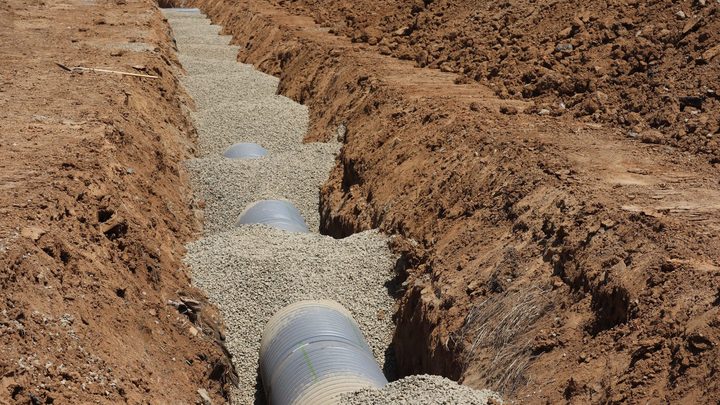 Vala aberta em rua asfaltada com tubulação de drenagem assentada no fundo, terra revolvida nas laterais e sinalização de obra ao redor.