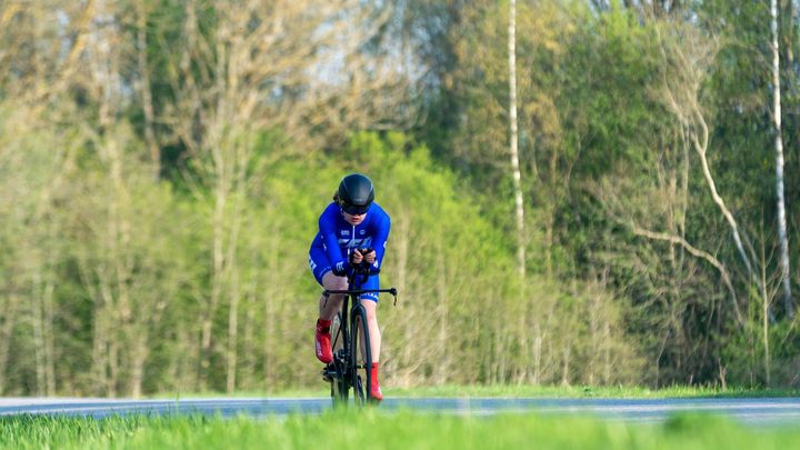 Ciclista em posição aerodinâmica sobre bicicleta de contrarrelógio, com capacete alongado, pedalando em estrada de campo aberto.
