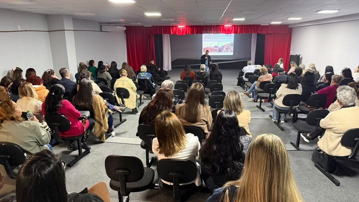 Auditório com plateia sentada de costas assistindo a uma apresentação, com um homem falando junto à tela projetada no palco de cortina vermelha.