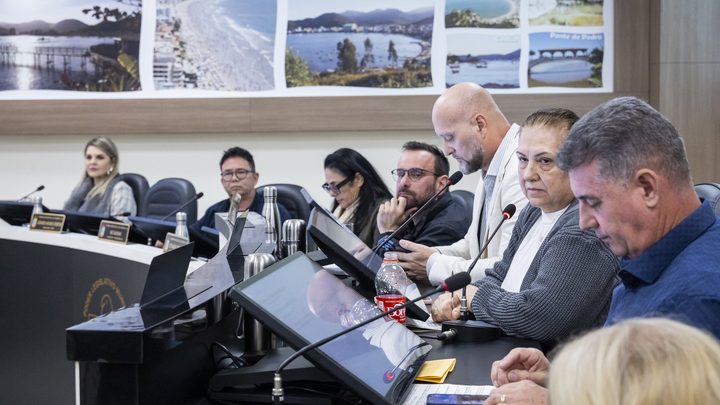 Vereadores sentados lado a lado na bancada do plenário da Câmara de Itapema durante sessão, com microfones e monitores à frente e um painel de fotos turísticas da cidade na parede ao fundo.